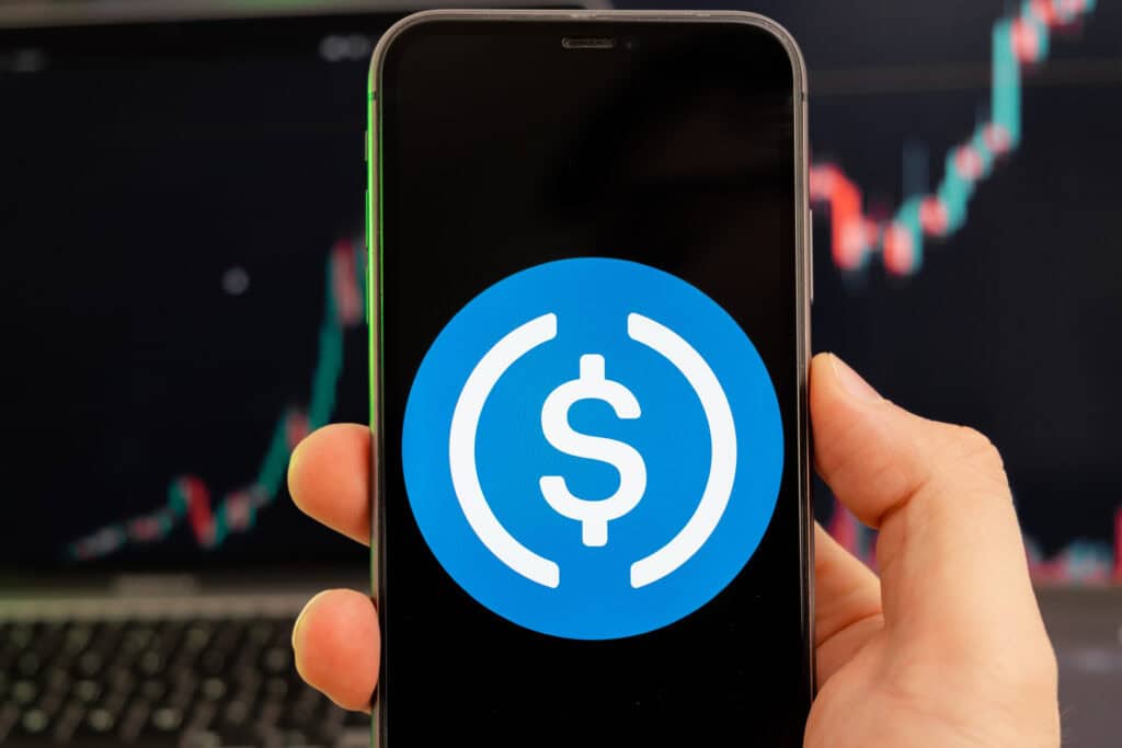 USD Coin cryptocurrency logo on the screen of a smartphone in mans hand with a growing trend on the chart on a green background.